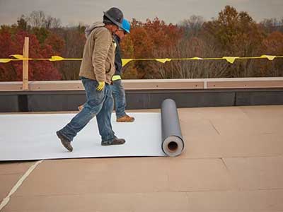 Two workers rolling out a white, singly-ply TPO sheet on a commercial roof